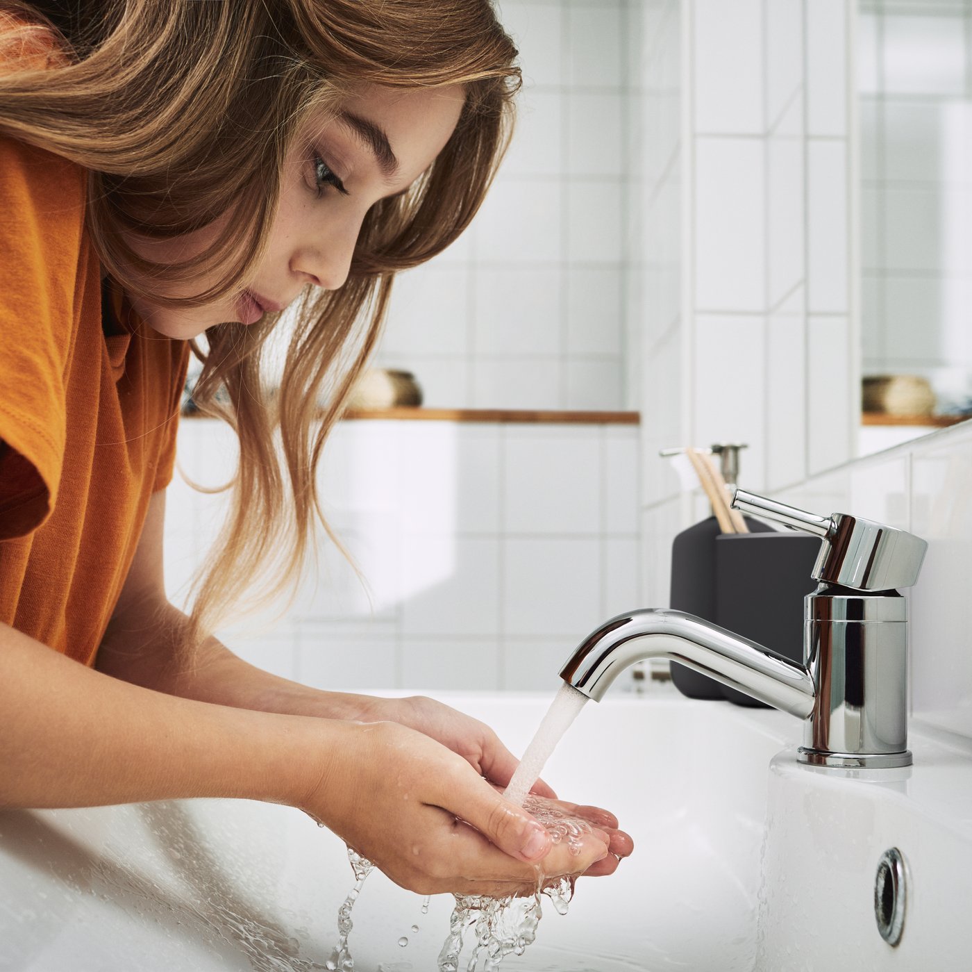 A girl is leaning over a sink in the bathroom washing her hands under the running water of a tap. 