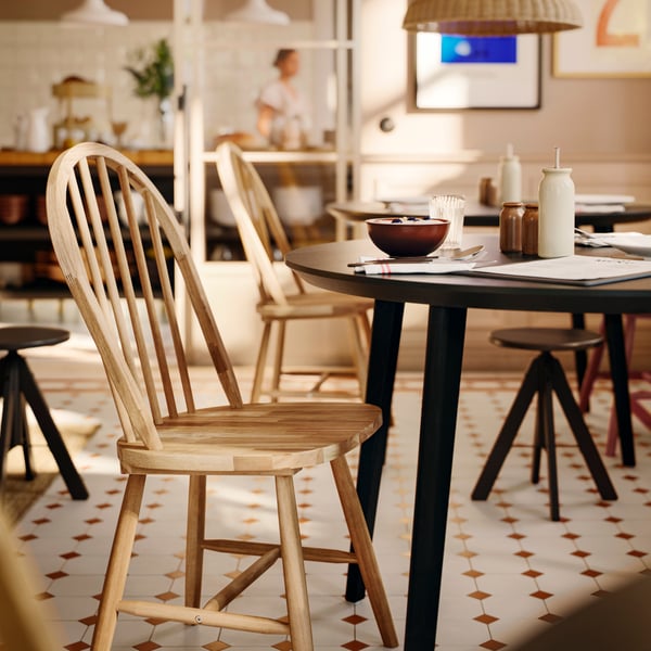 Une chaise SKOGSTA en acacia massif avec un dossier arqué et des fuseaux se trouve à une table ronde sombre dans une salle à manger commerciale.