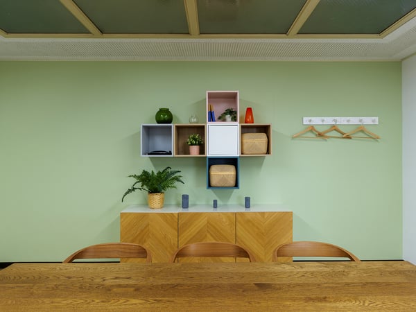 淡いグリーンの壁に飾り棚と小物を配置し、木製カウンターとスツールを合わせたナチュラルなワークスペース。
