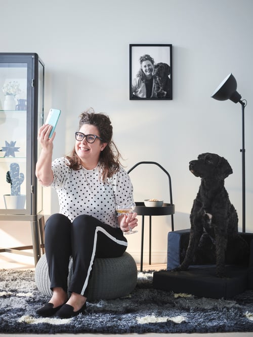 Living room with pouffe, side table, bed for pets, glass door cabinet, uplighter, frame, a person and a dog sitting down.