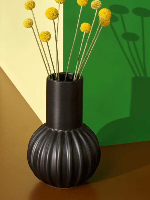 A black SKOGSTUNDRA vase with yellow flowers stands on a brown surface. It casts a shadow on the green wall behind it.