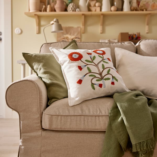 Närbild av beige EKTORP soffa med broderade blommiga kuddar och gröna prydnadskuddar, placerad framför hyllor med keramik.