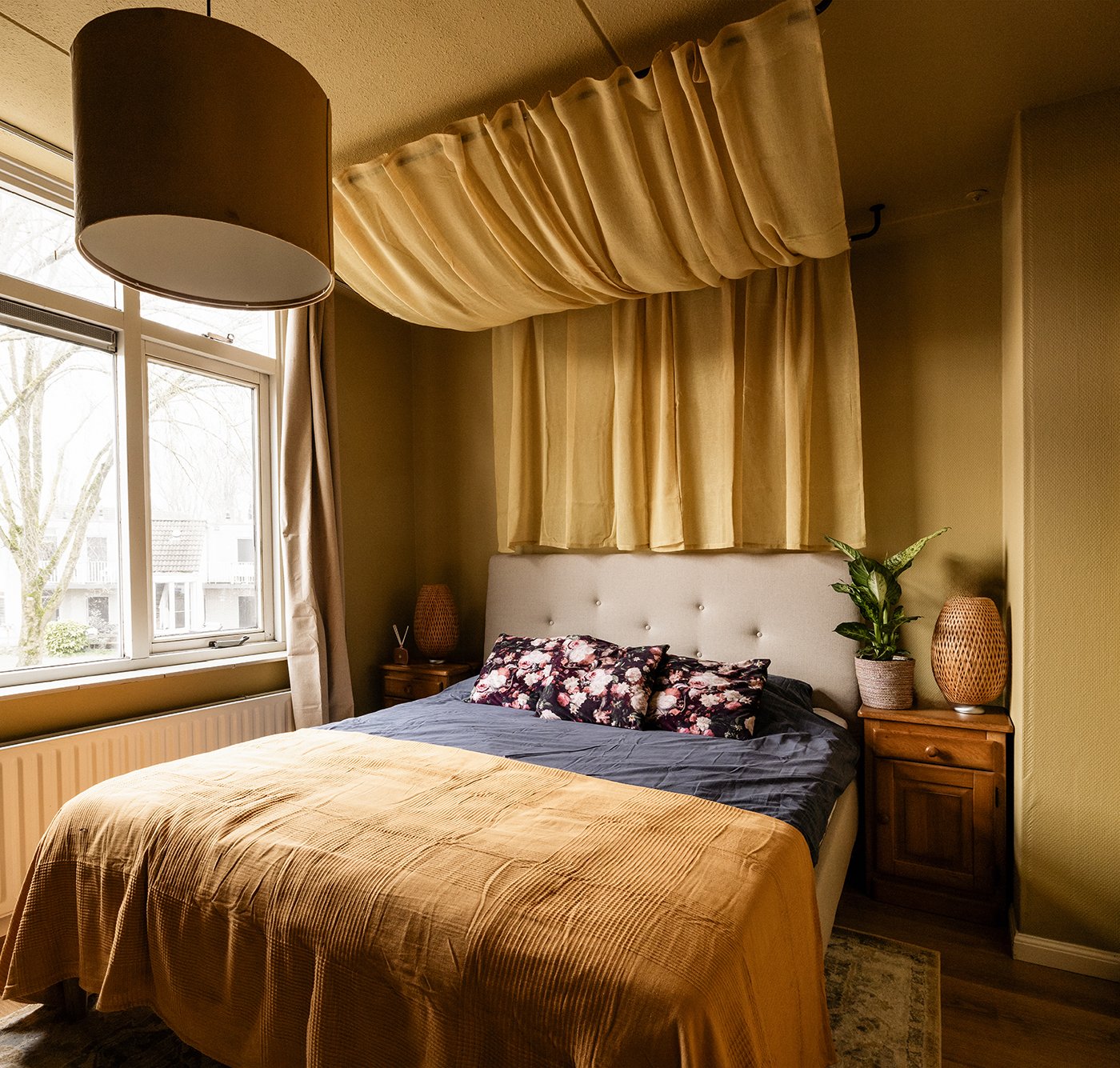 Slaapkamer met okergele muren. Hierin staat een Naggen beige IDANÄS bed, waarboven een okergeel gordijn hangt. Op het bed ligt een okergeel INDIRA sprei.