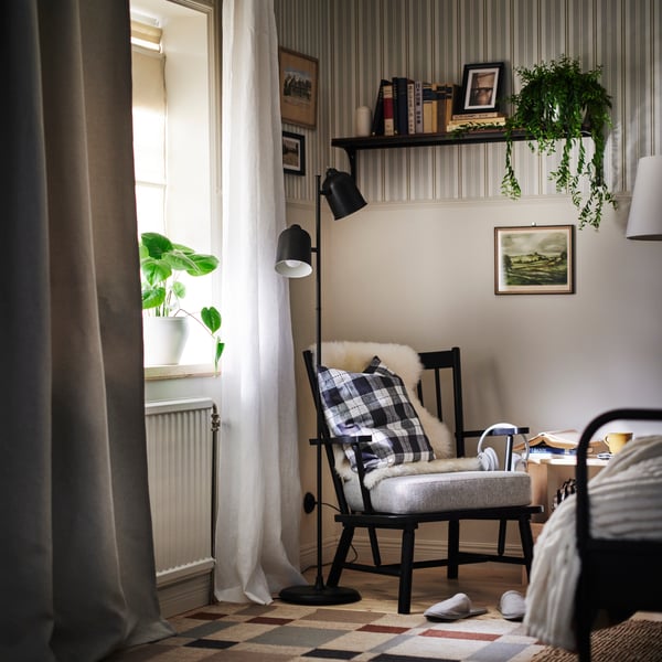 A corner of a bedroom with focus on PERSBOL armchair and KLÖVING floor lamp in black. Window to the left and bed at right.