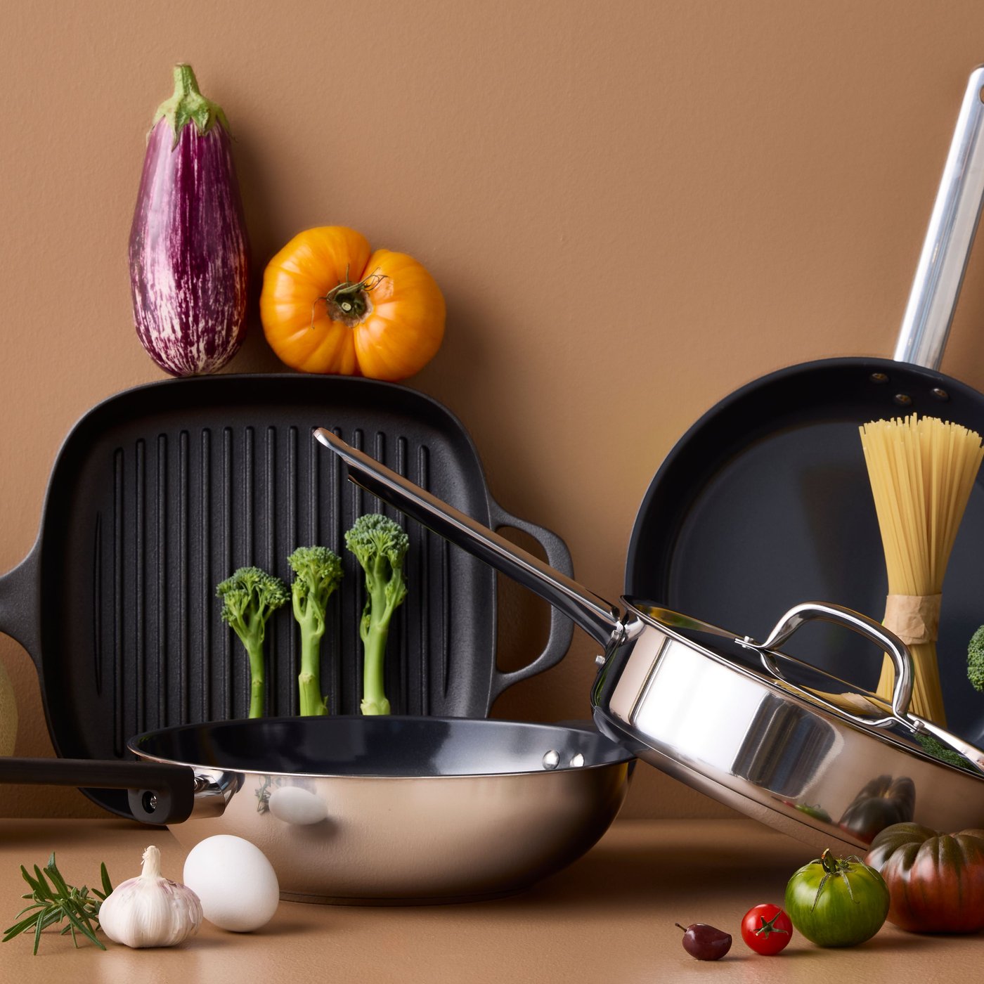 Four pans of different styles and materials are placed on a brown surface with different kinds of vegetables around them.