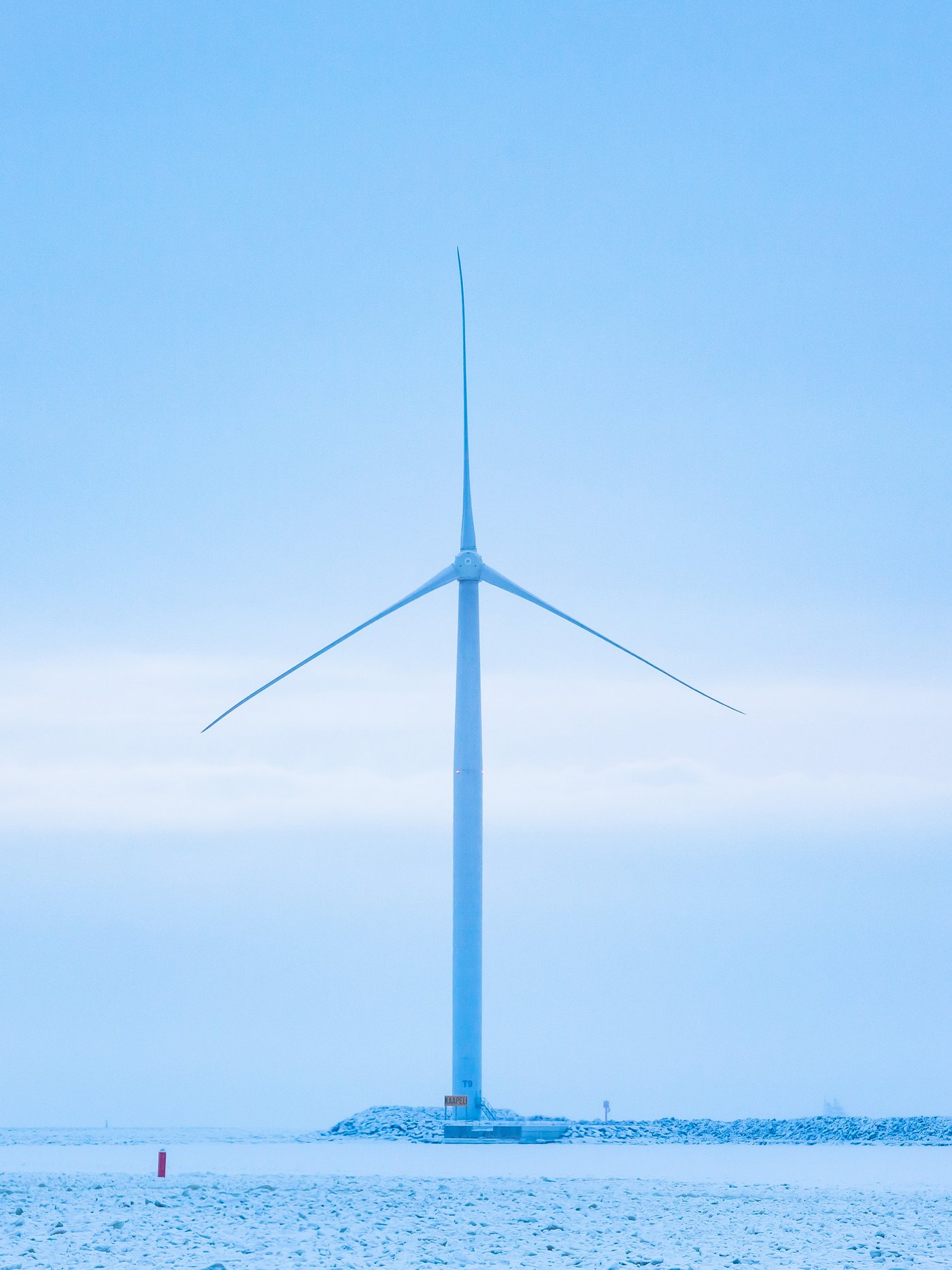 A single wind turbine at the end of a stone breakwater. The sky is grey blue with a single band of light grey cloud.