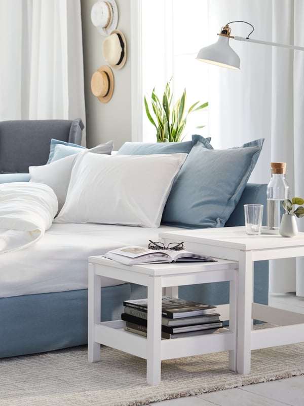 An IKEA HOLMSUND sofa-bed set up as a bed with pillows and covers, near a nest of HAVSTA white tables.