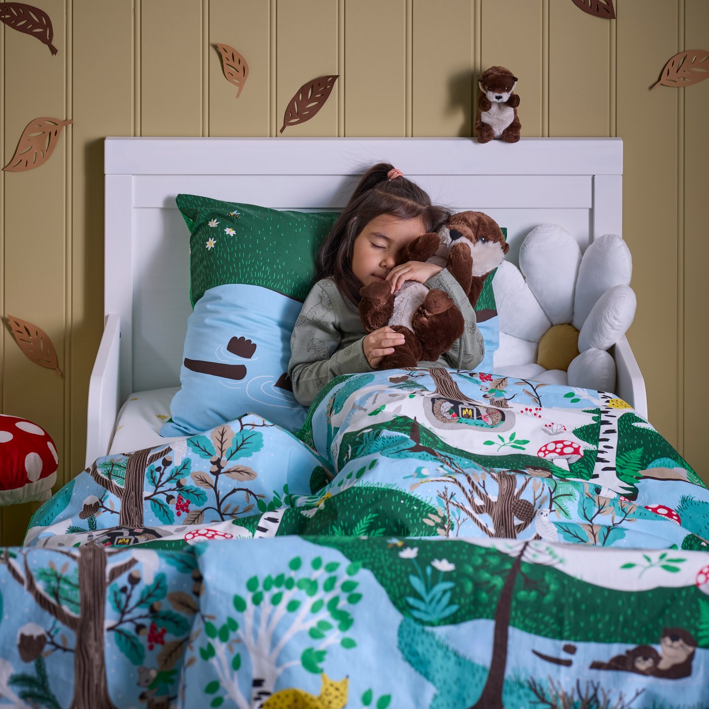 A child is lying in a white bed with SKOGSDUVA bed linen. The child is holding a SKOGSDUVA soft toy otter tightly.