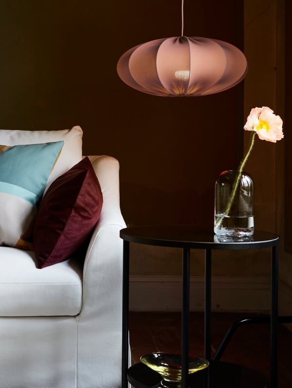 A living room with a hanging lamp, black table with flower in a glass jar and the corner of a white sofa with cushions.