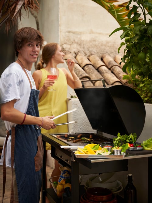 În aer liber, înfățișând o persoană care poartă un șorț GRILLTIDER albastru din bumbac, care frige mâncare la grătar alături de o altă persoană.