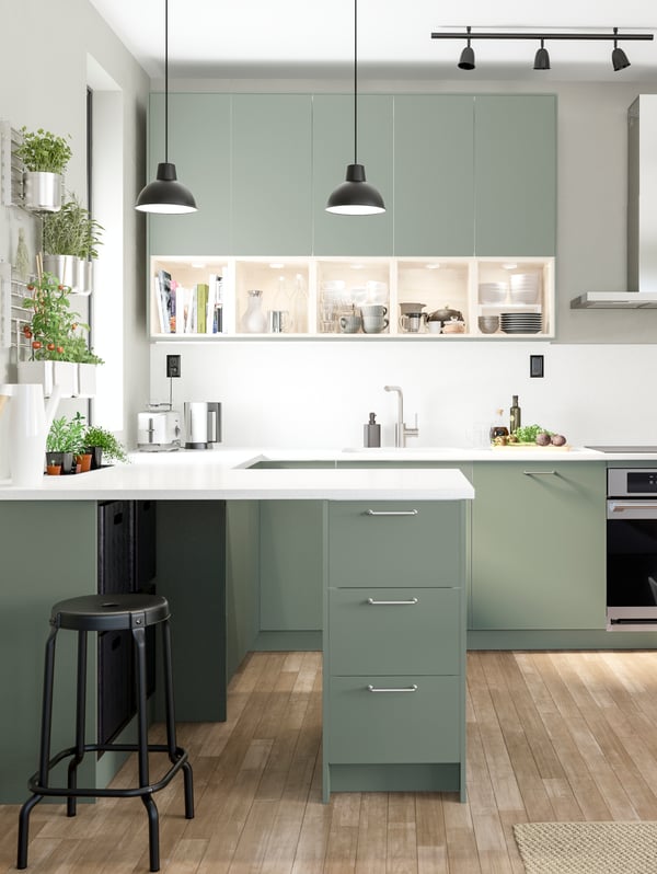 A grey-green kitchen with a white worktop, black pendant lamps and herbs in containers hanging on wall racks.