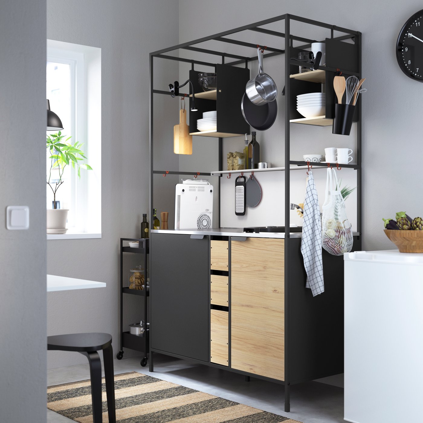 En modern köksuppsättning med en svart metallramshylla. Hyllan har träskåp och lådor, öppna hyllor med olika köksartiklar och en bänkskiva med en kaffebryggare och andra apparater. Det finns ett fönster på vänster sida som släpper in naturligt ljus, och golvet har en randig matta.