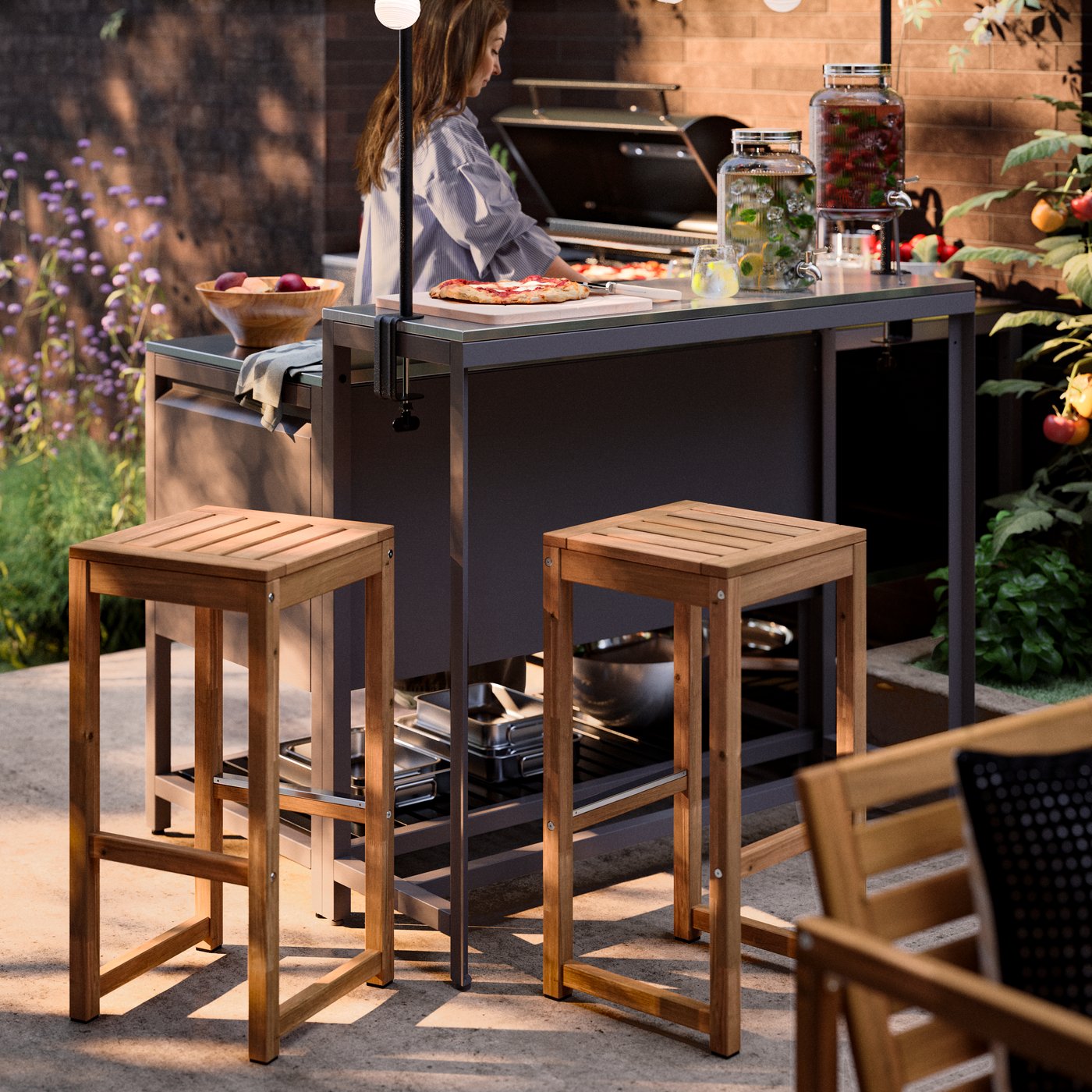 Two NÄMMARÖ bar stools stand beside an outdoor kitchen on a terrace where a person is cooking.