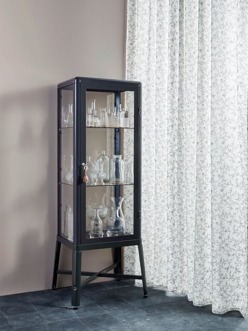 Le coin d’un salon avec des rideaux blancs, un mur couleur crème clair et une vitrine avec différents objets en verre.