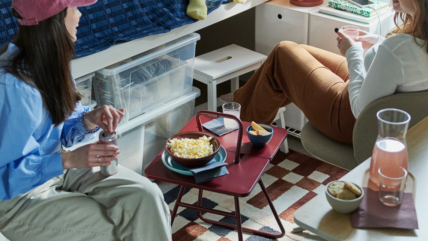 Three students in a dorm room gathered around an IKEA PS 2026 side table, with a KULGLASS speaker lamp in the background.