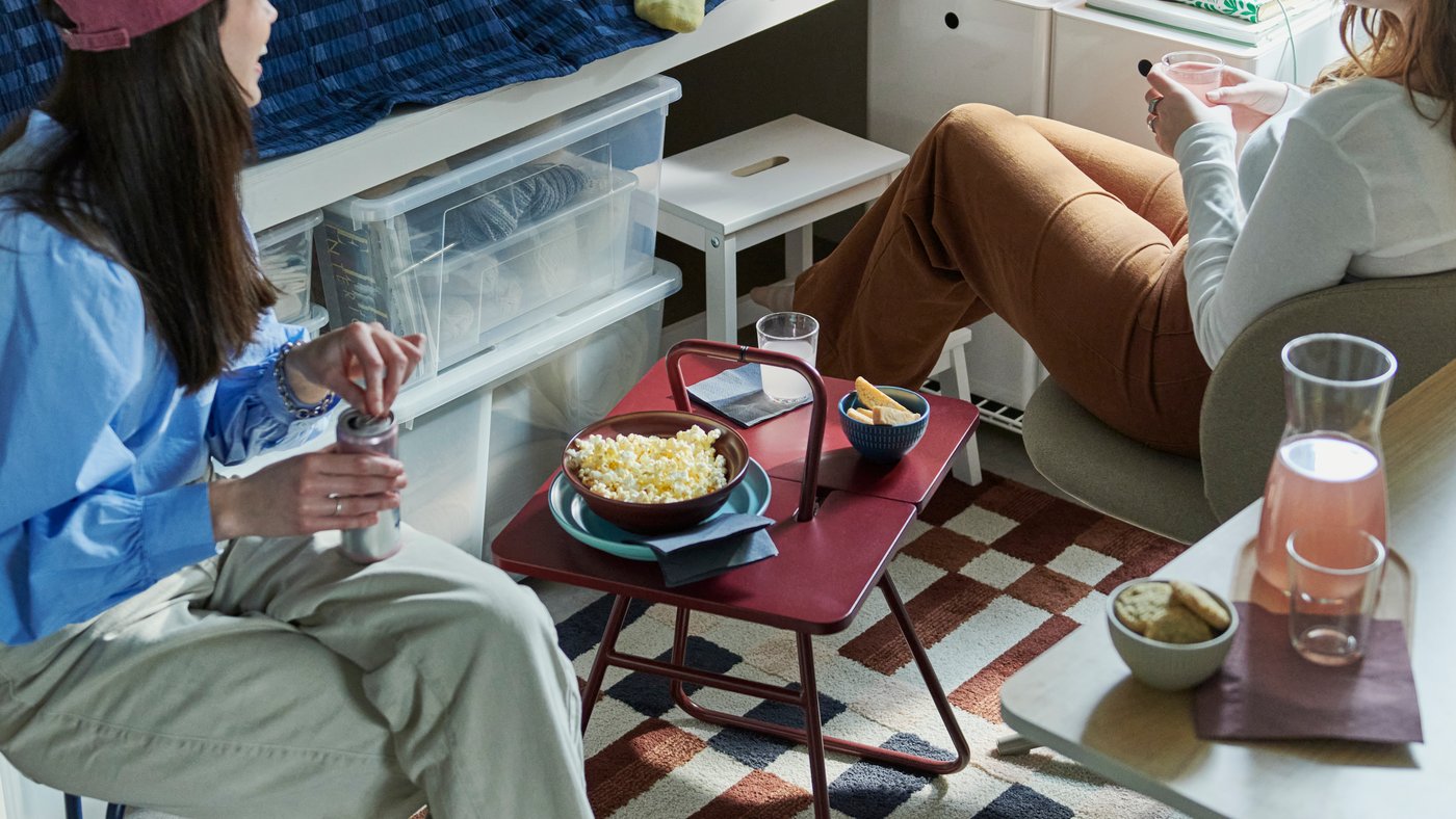 Three students in a dorm room gathered around an IKEA PS 2026 side table, with a KULGLASS speaker lamp in the background.