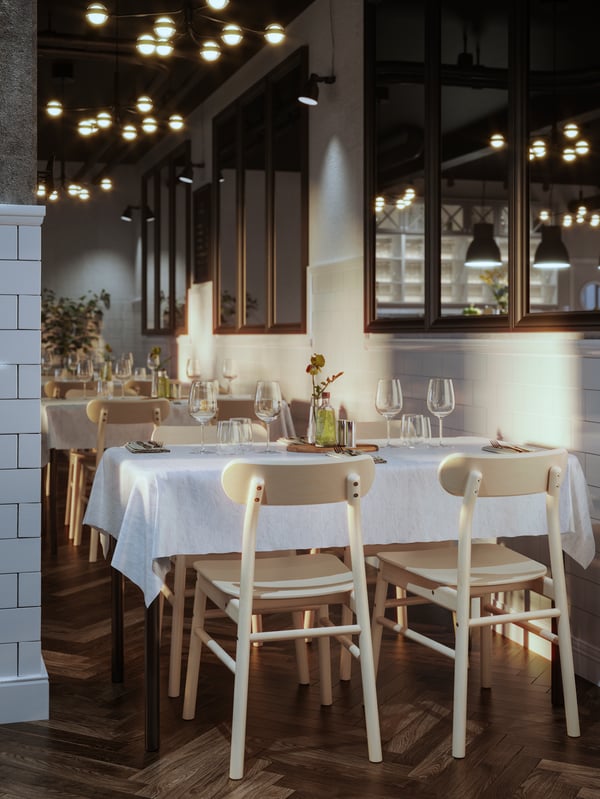 Ein TOMMARYD Tisch steht in einem sanft beleuchteten Restaurant. Um ihn herum befinden sich vier Holzstühle. Er ist mit einer weißen Tischdecke und Gläsern gedeckt.