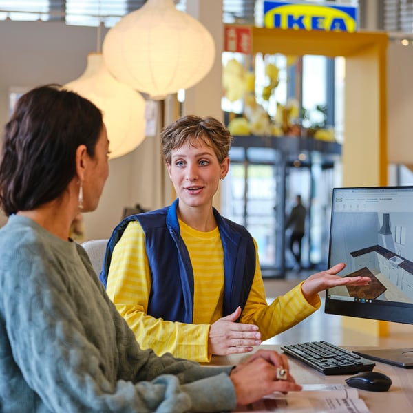 Una co-worker IKEA for Business in divisa blu e gialla seduta davanti al PC che spiega un progetto a una cliente con partita IVA, vestita con un pullover verde.