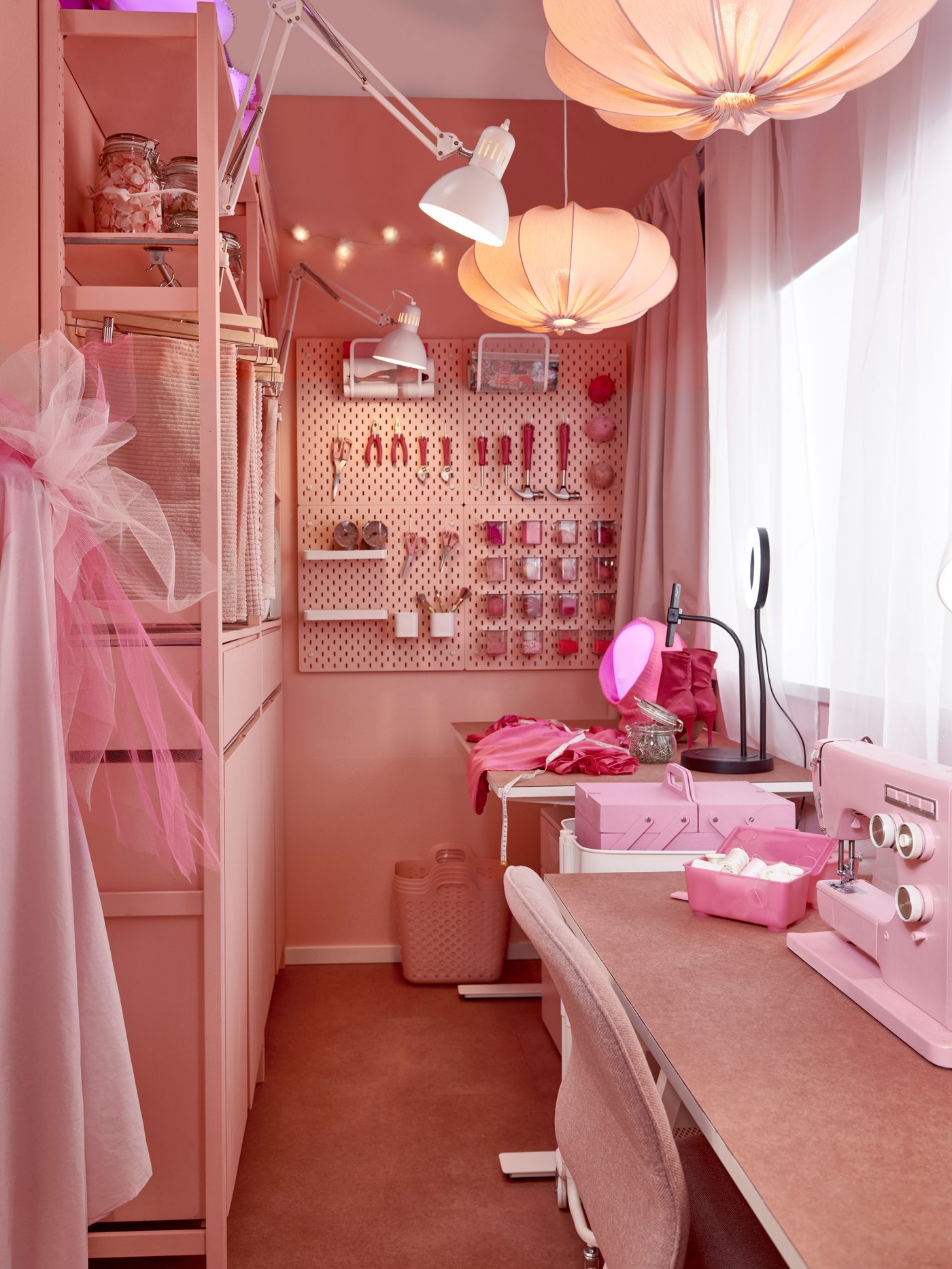 A room with all the furniture and accessories in pink. A desk  with a chair is by the window to the right and a shelving unit to the left and back of the image.