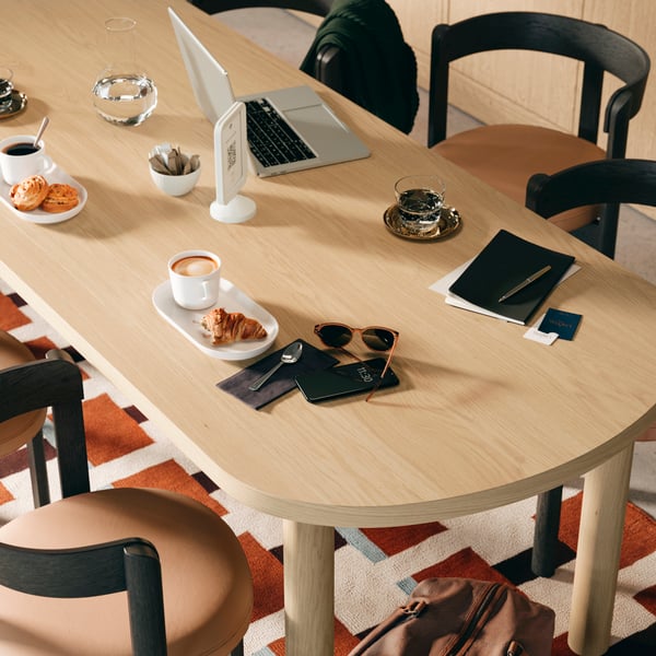 A close-up of a STOCKHOLM 2025 table in oak veneer with 6 dark brown chairs, arranged with coffee and pastries.