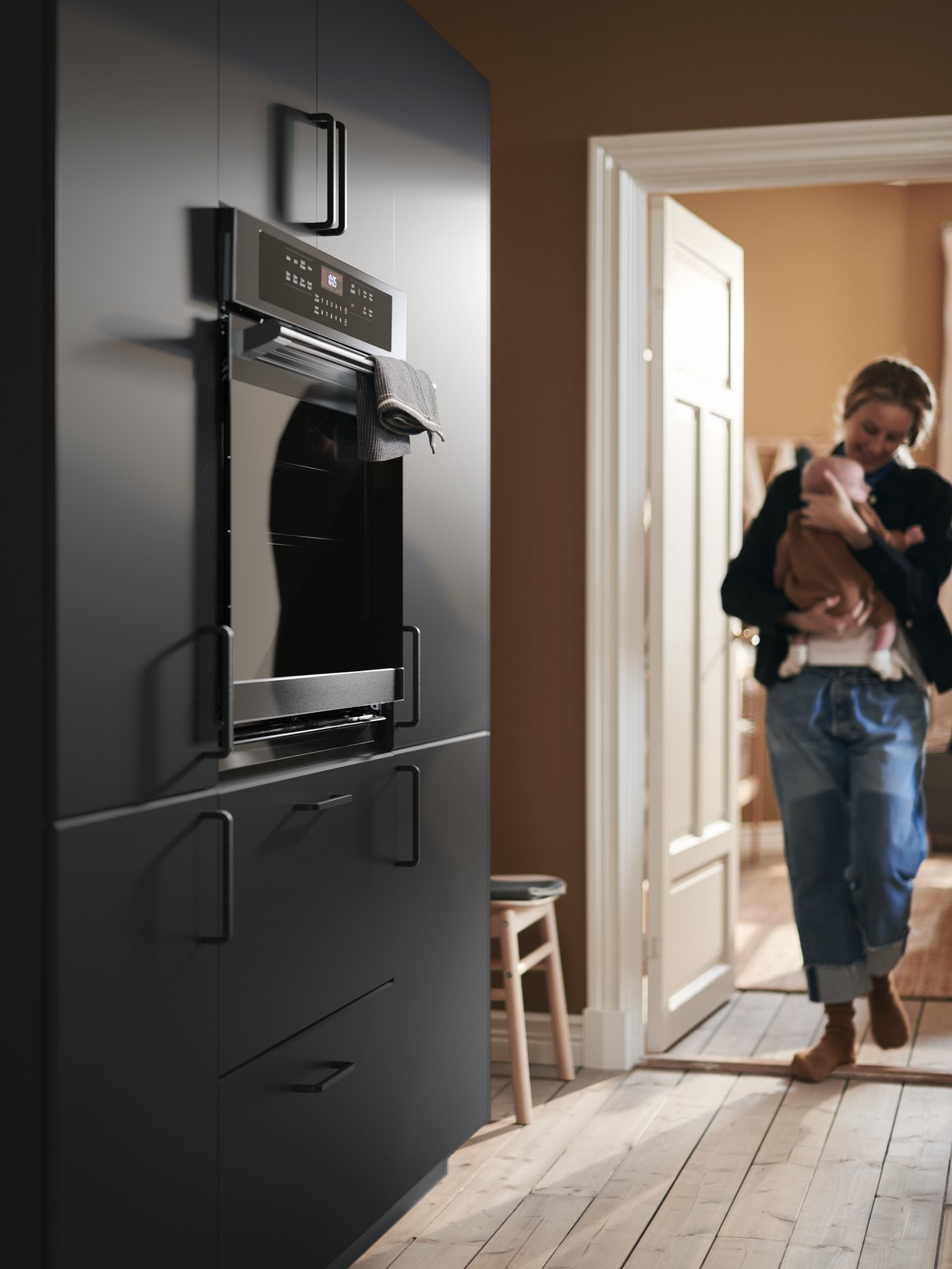 A mother cradles her baby as she enters the kitchen, approaching a wall of cabinets with KUNGSBACKA fronts.
