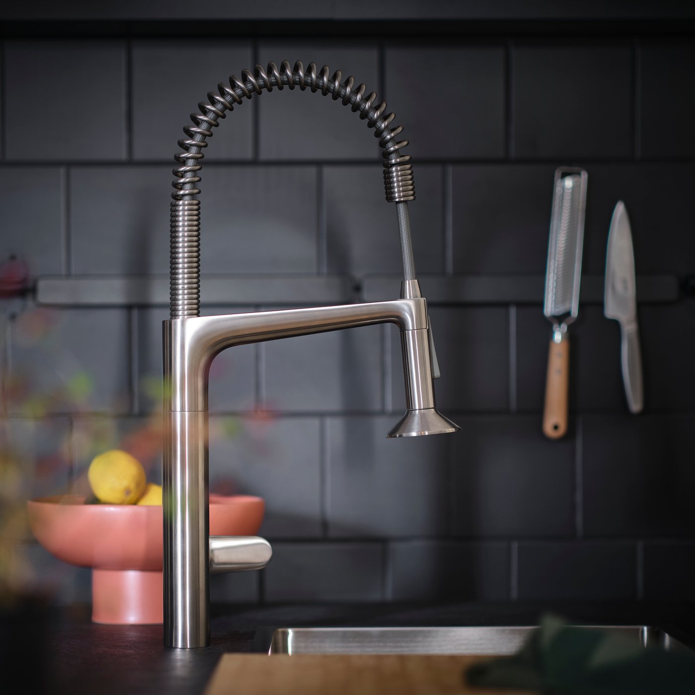 Et MAJSJÖN køkkenblandingsbatteri med hane i rustfrit stål trækkes ned med sprøjtestråle i profil ved en mørk flisebelagt væg og en skål citroner.