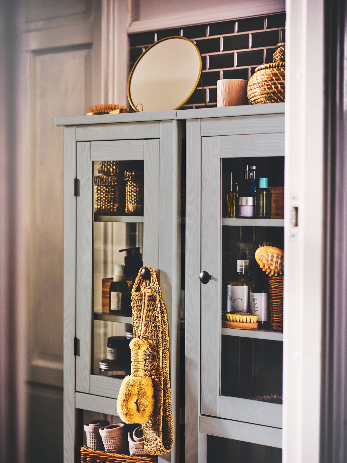 Two grey HEMNES high cabinets with glass-doors, toiletries placed inside, boxes with lids and a mirror on top.