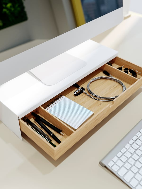 Escritorio con un soporte ELLOVEN para pantalla de computador con un cajón abierto de bambú que contiene un cable, una libreta y lápices ordenados. 