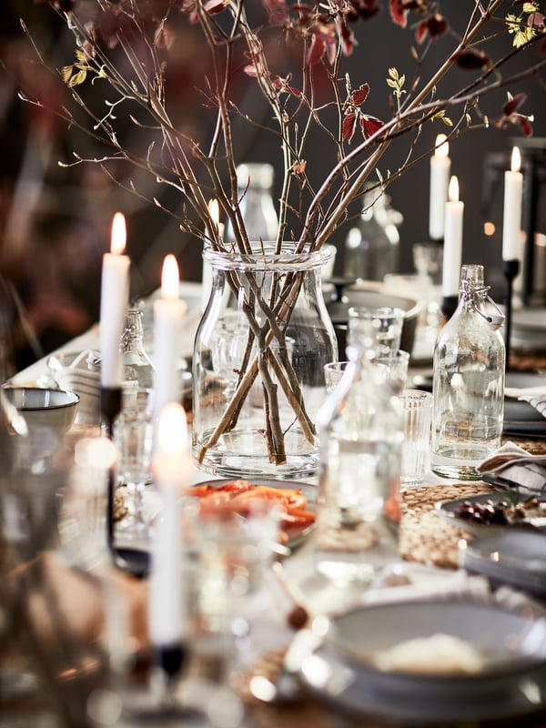 Stimmungsvoll gedeckter Tisch mit Kerzen, Glasvase mit Zweigen und schlichten Tellern – warm, herbstlich.