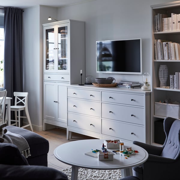 A white and grey tone living room with an IDANÄS glass door cabinet, chest of drawers and bookcase in a line along the wall.