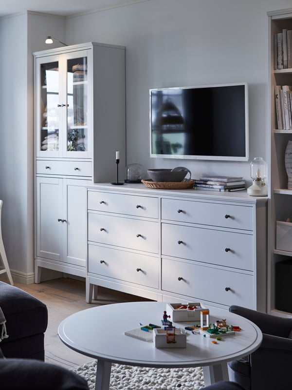 Un salon dans les tons blancs et gris avec une vitrine IDANÄS, une commode et une bibliothèque le long du mur.