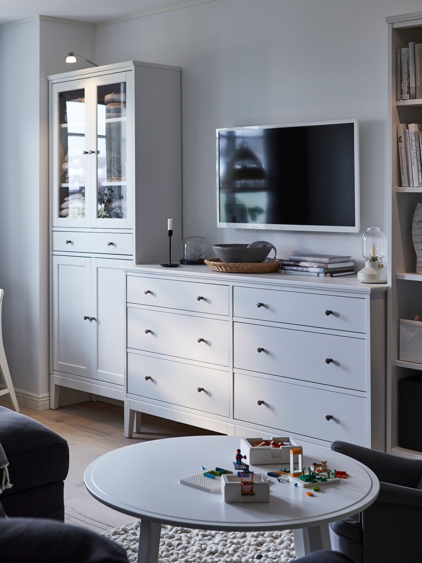 A white and grey tone living room with an IDANÄS glass door cabinet, chest of drawers and bookcase in a line along the wall.