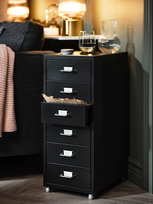 A black HELMER drawer unit on castors stands at an angle next to a sofa. Two lit brass lamps are on a shelf behind the sofa.