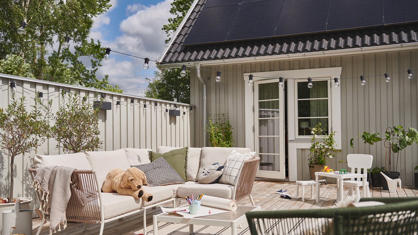 En uteplats med ljusa trämöbler, kuddar och filtar, leksaker och krukväxter framför ett hus med panelväggar och glasdörrar. Solpaneler syns på taket och träd i bakgrunden.