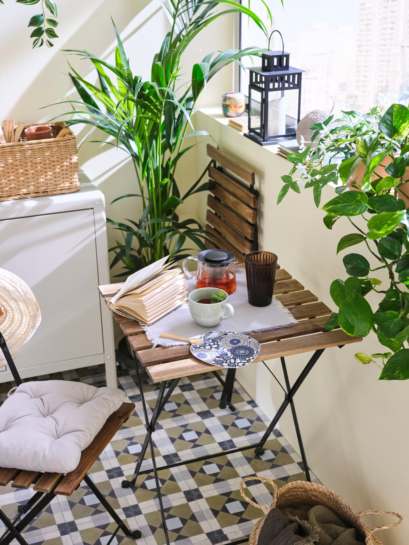 Black/light-brown stained acacia outdoor TÄRNÖ table and 2 chairs with seat cushions are in a plant-filled, enclosed balcony.