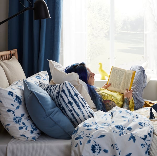 Poltrona em branco com almofada STJÄRNTULPAN em azul escuro e vermelho e uma manta POLARVIDE em azul, junto a uma grande janela.