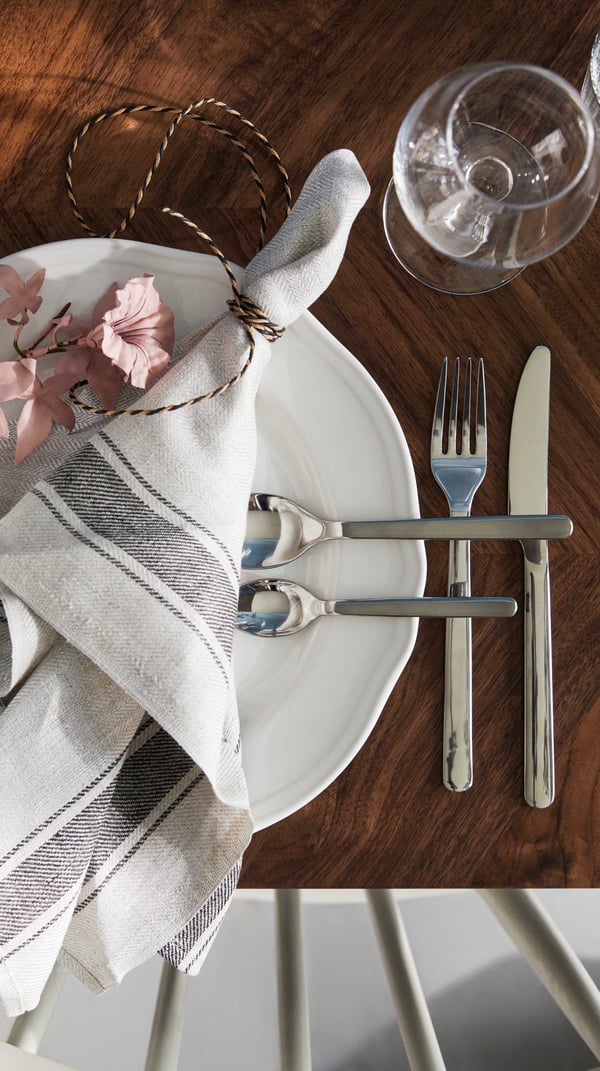 A white traditional plate with a white and grey fabric napkin rolled on top of it, with an IKEA 365+ steel cutlery set next to it and an empty glass of wine, all resting on a walnut table.