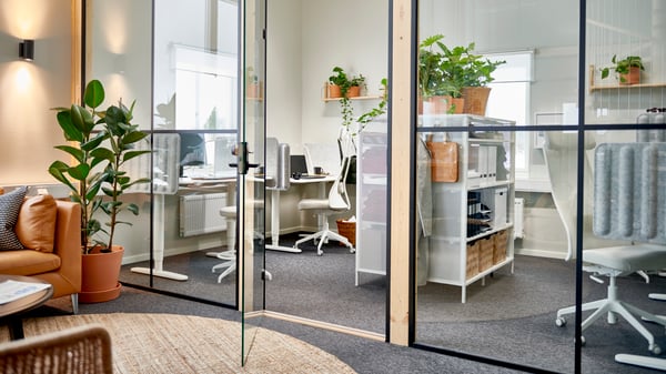 Modernt kontor med glasväggar, vita skrivbord med datorer, kontorsmaterial och krukväxter. Ergonomiska stolar, hyllor med förvaringskorgar och växter. Utanför kontorsytan finns en sittgrupp med brun lädersoffa, rund matta och stor krukväxt i ett ljust och inbjudande rum.