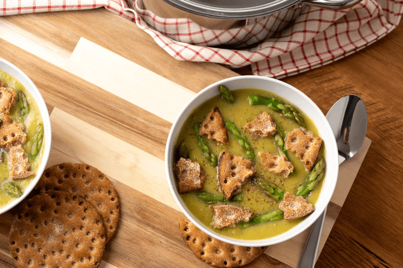 Eine Spargelsuppe mit Knäckebrot-Crunch auf einem Holztisch präsentiert.