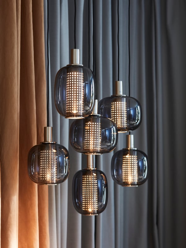 Two HÖGVIND pendant lamps with 3 lamps hang from the ceiling in front of some curtains. The lamps are lit.