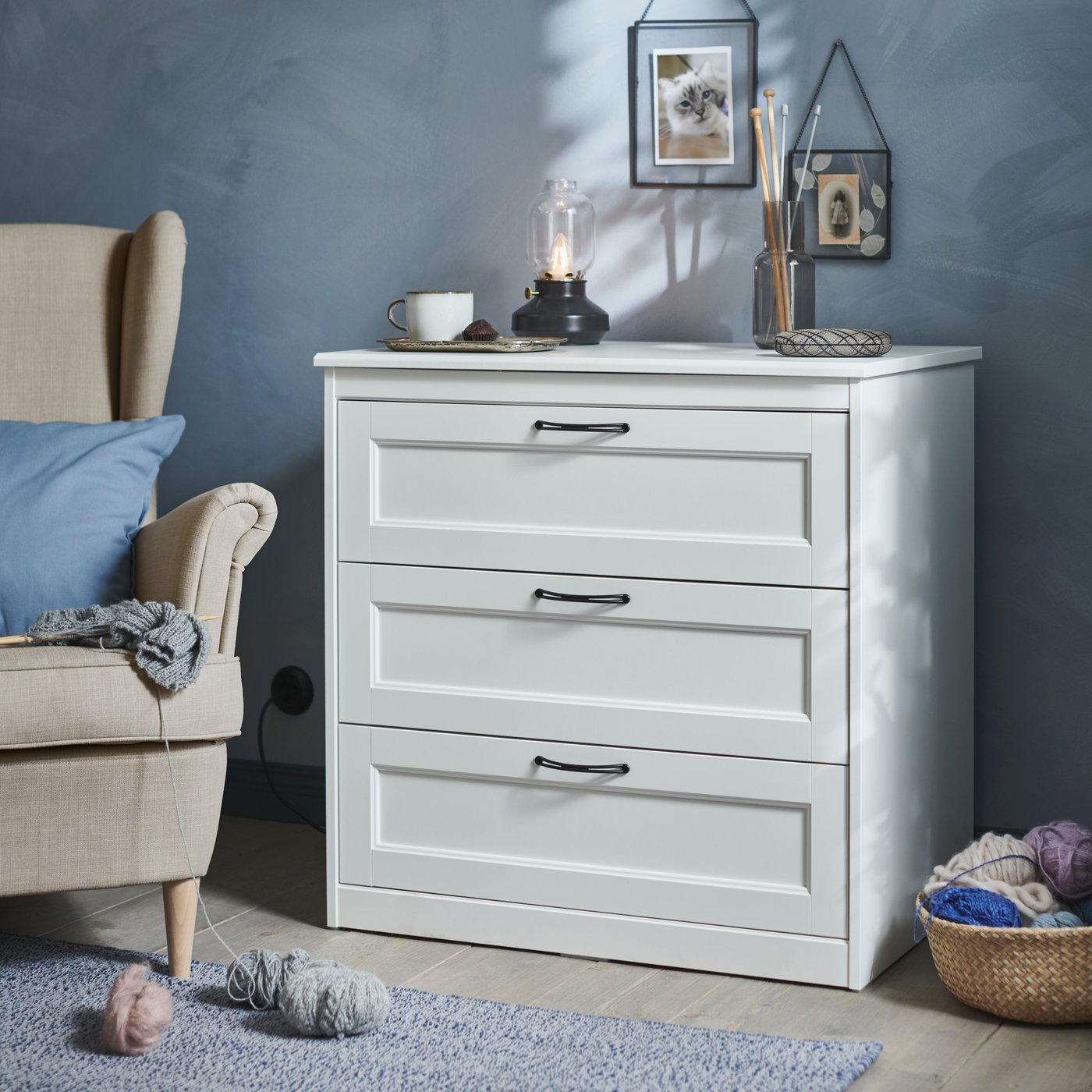 Vit SONGESAND byrå med tre lådor mot en blå vägg, dekorerad med lampa, kopp och små inredningsdetaljer, intill en fåtölj och väggdekorationer.