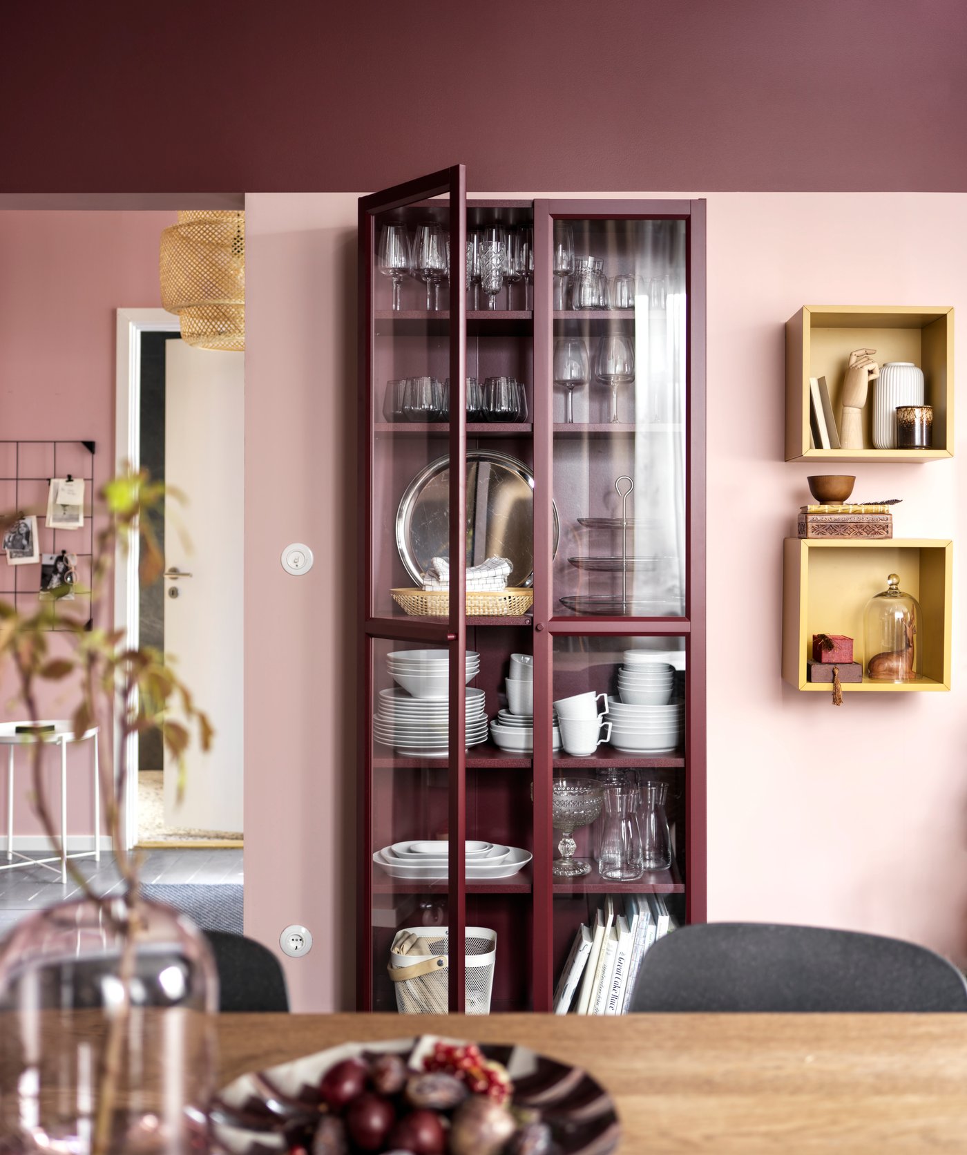 In primo piano c'è un tavolo di legno marrone con vasi e ciotole decorative. Sullo sfondo è esposta una vetrina rossa con piatti, bicchieri e decorazioni.