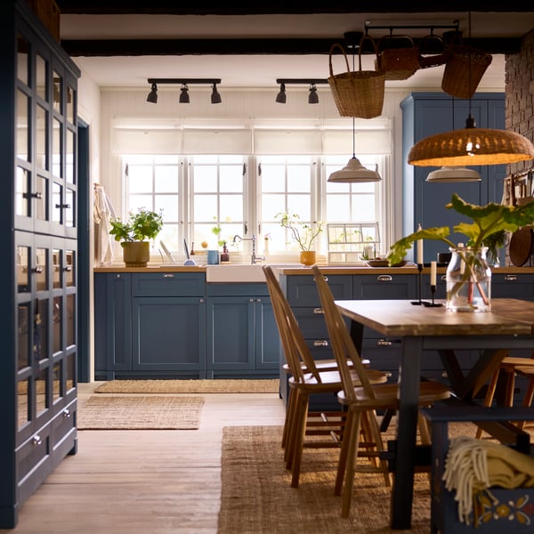 A METOD kitchen with fronts in LERHYTTAN blue has large windows, a kitchen island with drawers and a dining table.