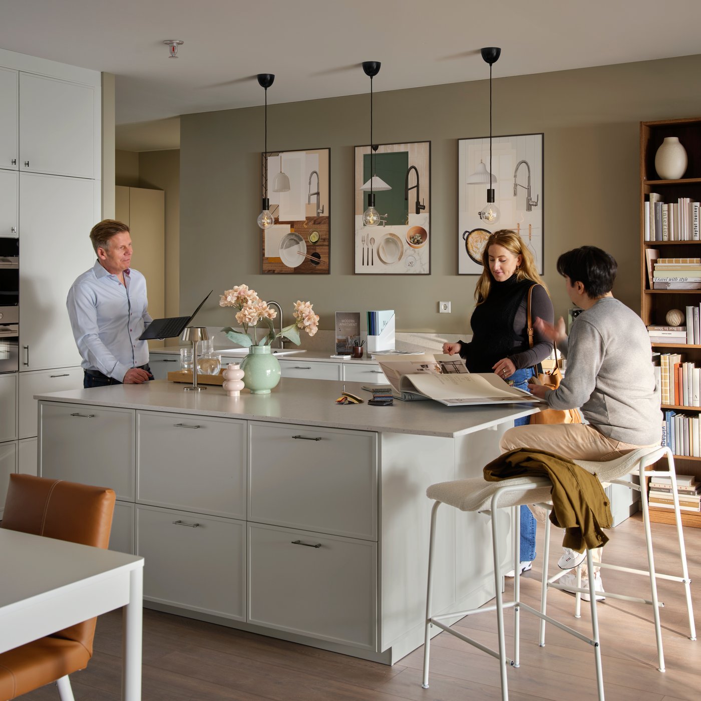 Cucina espositiva moderna bianca con grande isola centrale dove un venditore parla con una giovane coppia seduta su sgabelli alti.