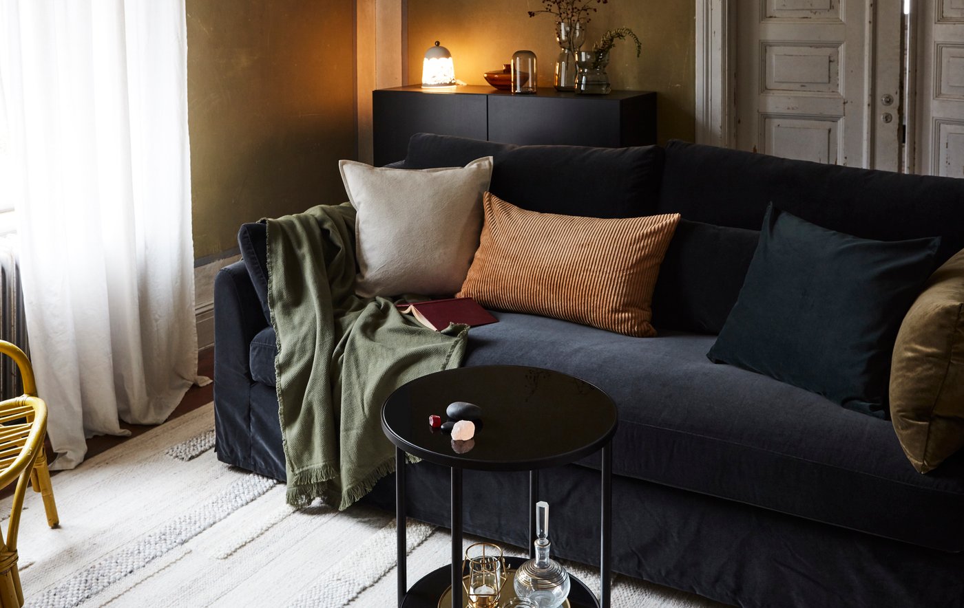 A living room with a sofa, cushions, throw, rug, a small table, a sideboard with a lamp, vases, and a rattan armchair.