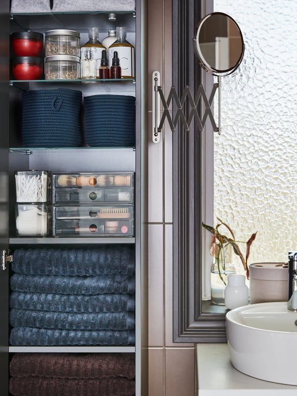 An open high cabinet with dark-grey towels, see-through boxes, jars and blue baskets on the shelves and a mirror on the side.