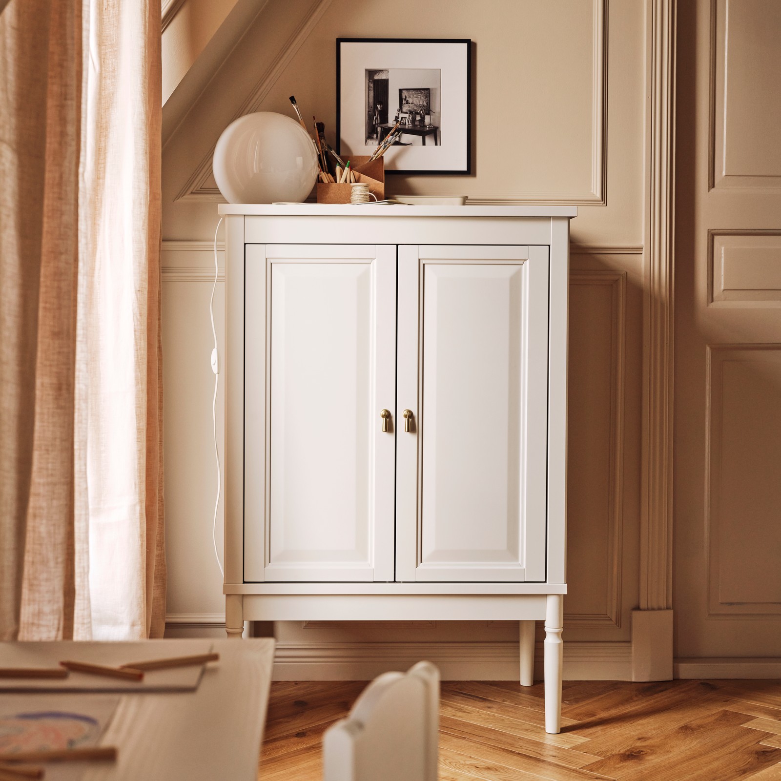 An off-white FÄRJKARL cabinet with a FADO table lamp on top stands against the wall near a window in a living room.