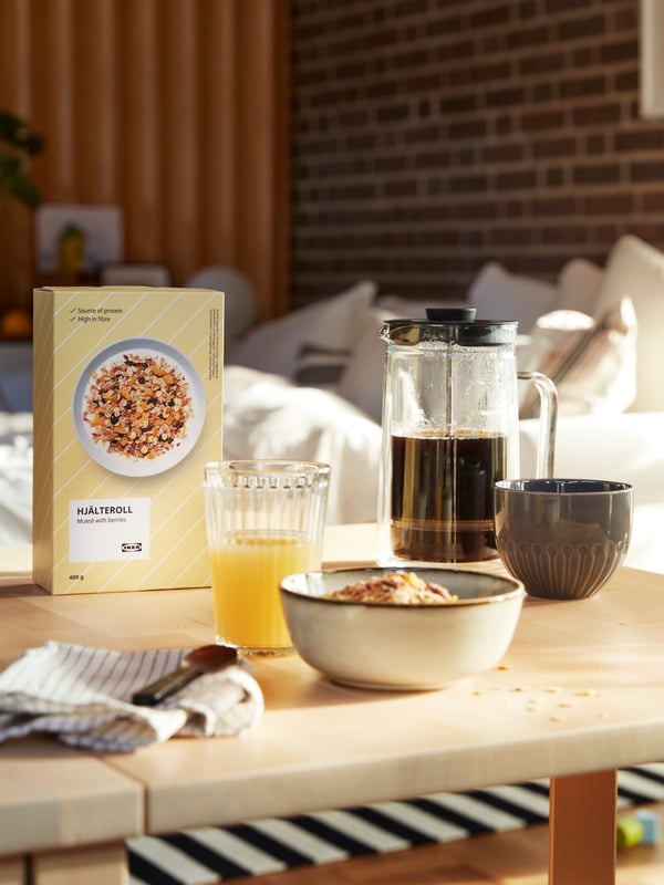 On a NORDEN birch-coloured table are: HJÄLTEROLL Muesli in a grey GLADELIG bowl, a box of HJÄLTEROLL Muesli (400 g) with dried berries, a cup of orange juice, an EGENTLIG coffee/tea maker filled with coffee, and a grey STRIMMING mug. A bed is in the background. 