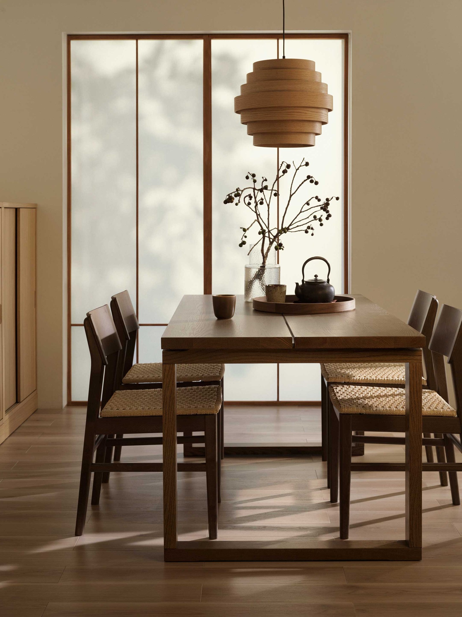 A warm and inviting dining room, perfect for gathering with loved ones. A sturdy, rectangular wooden dining table takes centre stage, surrounded by four matching wooden chairs with comfortable woven seats. Above the table, a unique, layered wooden pendant light adds a touch of natural elegance. On the table, a clear glass vase holds a branch with berries, bringing a hint of the outdoors in. A dark teapot and small cups are neatly arranged on a wooden tray, ready for a cosy tea time. In the background, a screen with translucent panels allows soft, diffused light to fill the room, creating a serene and harmonious atmosphere. To the left, a glimpse of a wooden cabinet offers practical storage, completing this beautifully styled space.