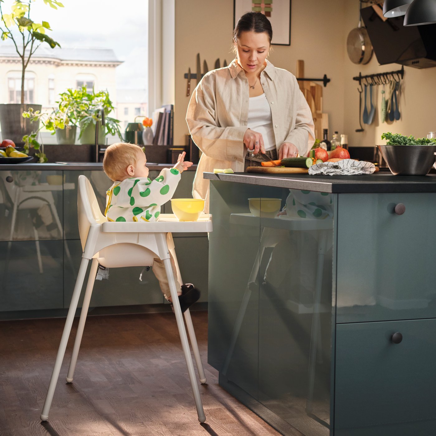 Ein Baby sitzt in einem ANTILOP-Hochstuhl neben einer Küchenarbeitsplatte, trägt ein weisss-grünes KLADDIG-Lätzchen mit langen Ärmeln und streckt die Arme aus.